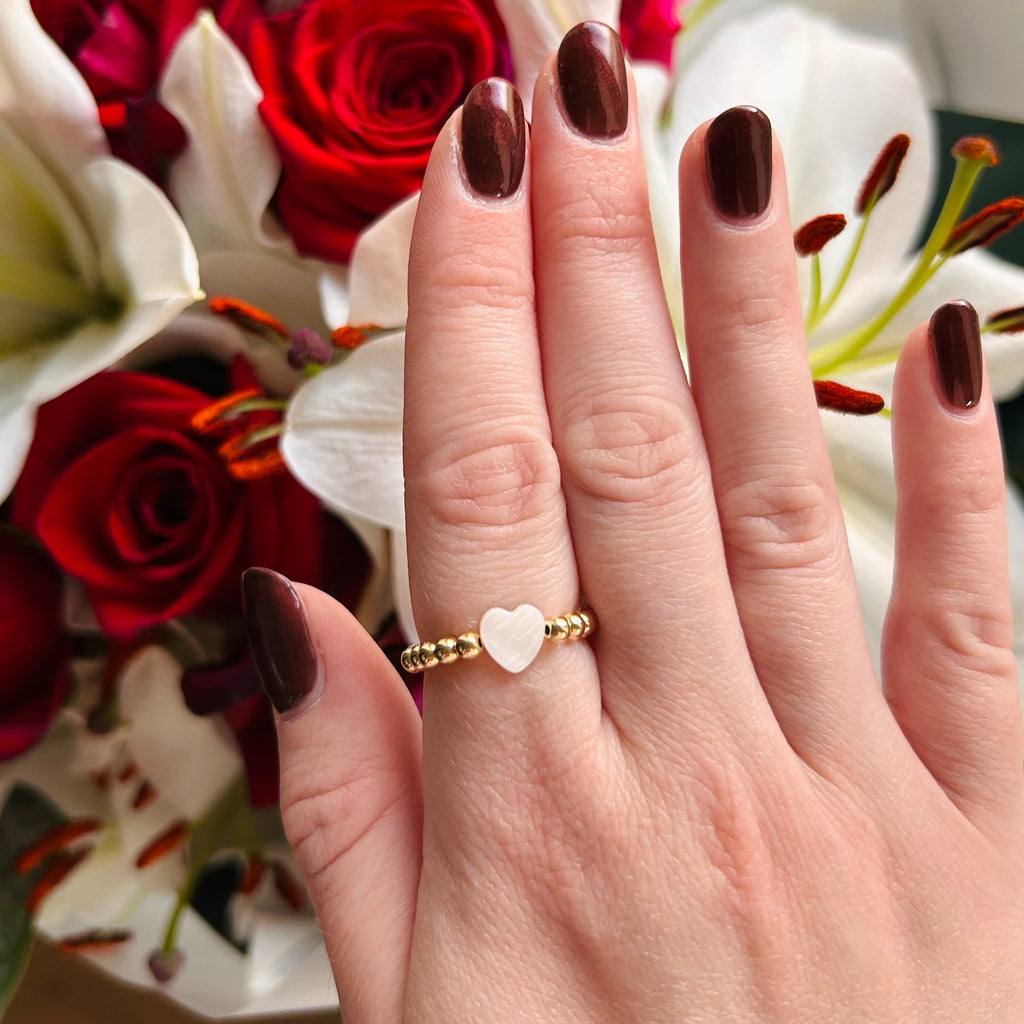 Hand wearing a gold ring with a heart design in front of flowers