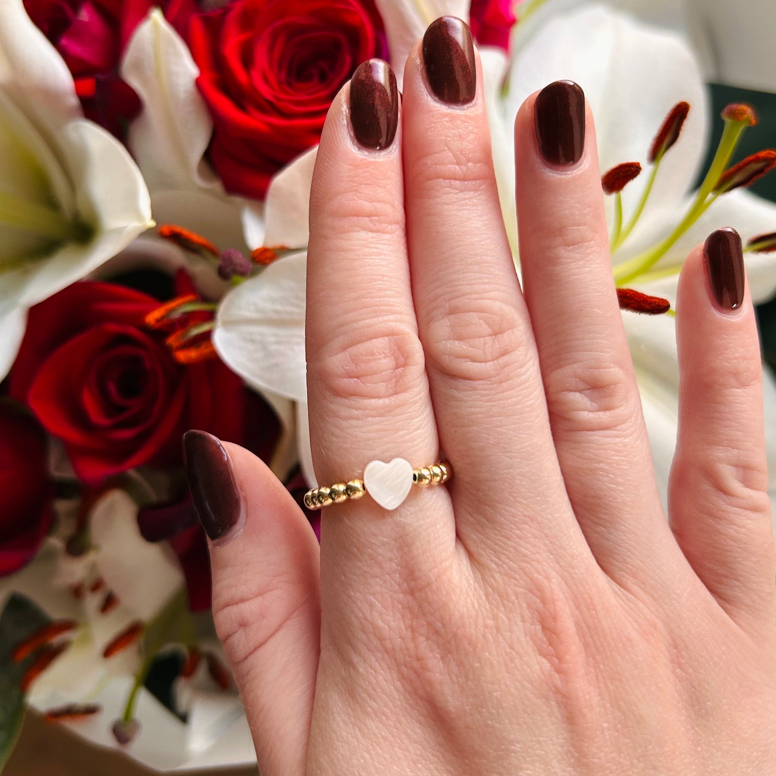 Hand wearing a gold ring with a heart design in front of flowers